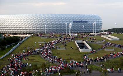 A photograph of a large football stadium in the background with crowds of people walking up to it in the foreground.