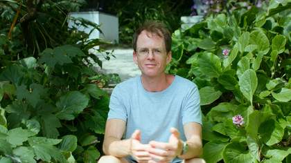 A person wearing a white t-shirt and glasses sitting among many green plants
