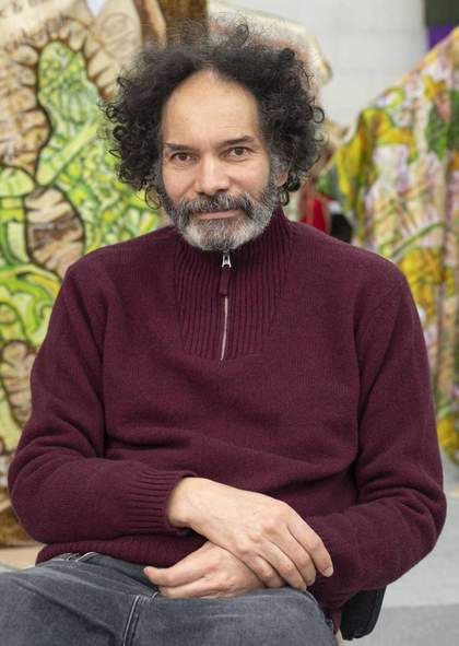 The artist Hew Locke wearing a purple top and sat in a chair.