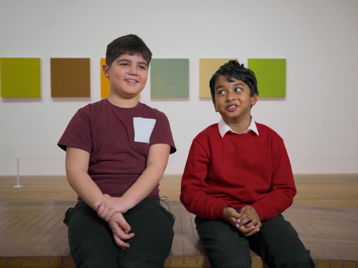 Two boys with school uniform on look at the camera, sitting on a bench in the gallery
