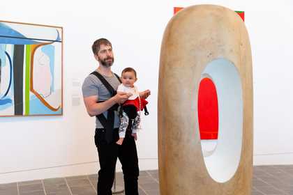 a man holding a baby in a sling looks at a big Barbara Hepworth sculpture in a gallery space