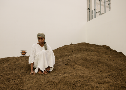 The artist sits on a large pile of earth inside what looks like a contemporary art space. They wear a white dress and leopard print headwrap.